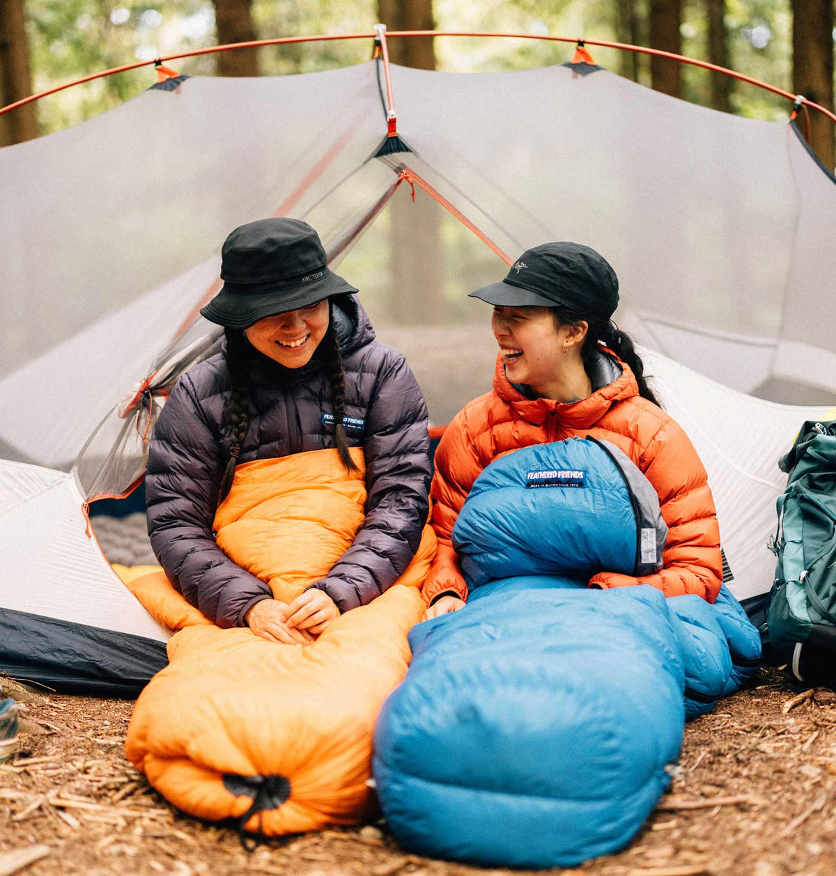 Two women in their tangerine and azure colored sleeping bags at camp in the forest