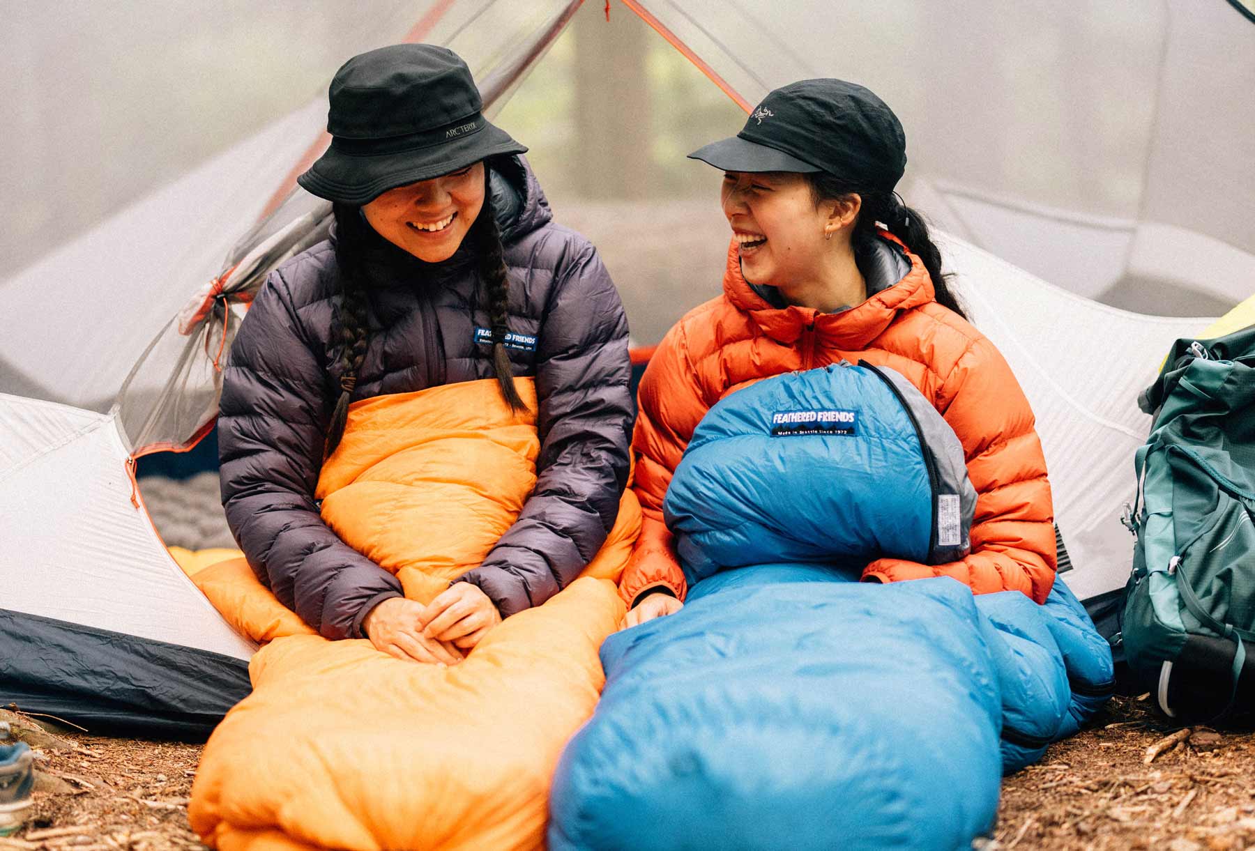Two women in their tangerine and azure colored sleeping bags at camp in the forest