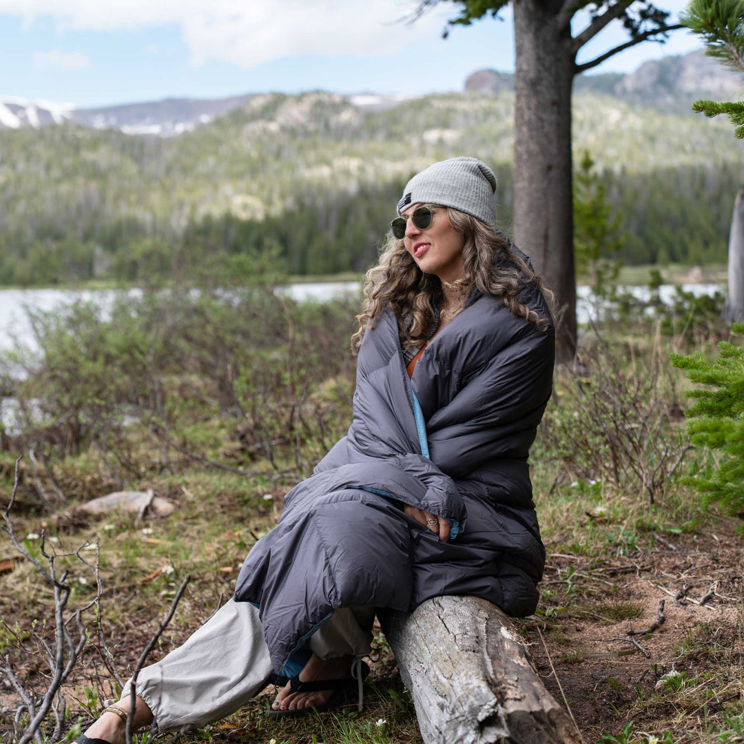 Person wrapped in a Feathered Friends grey lightweight down blanket by a lake with trees in the background