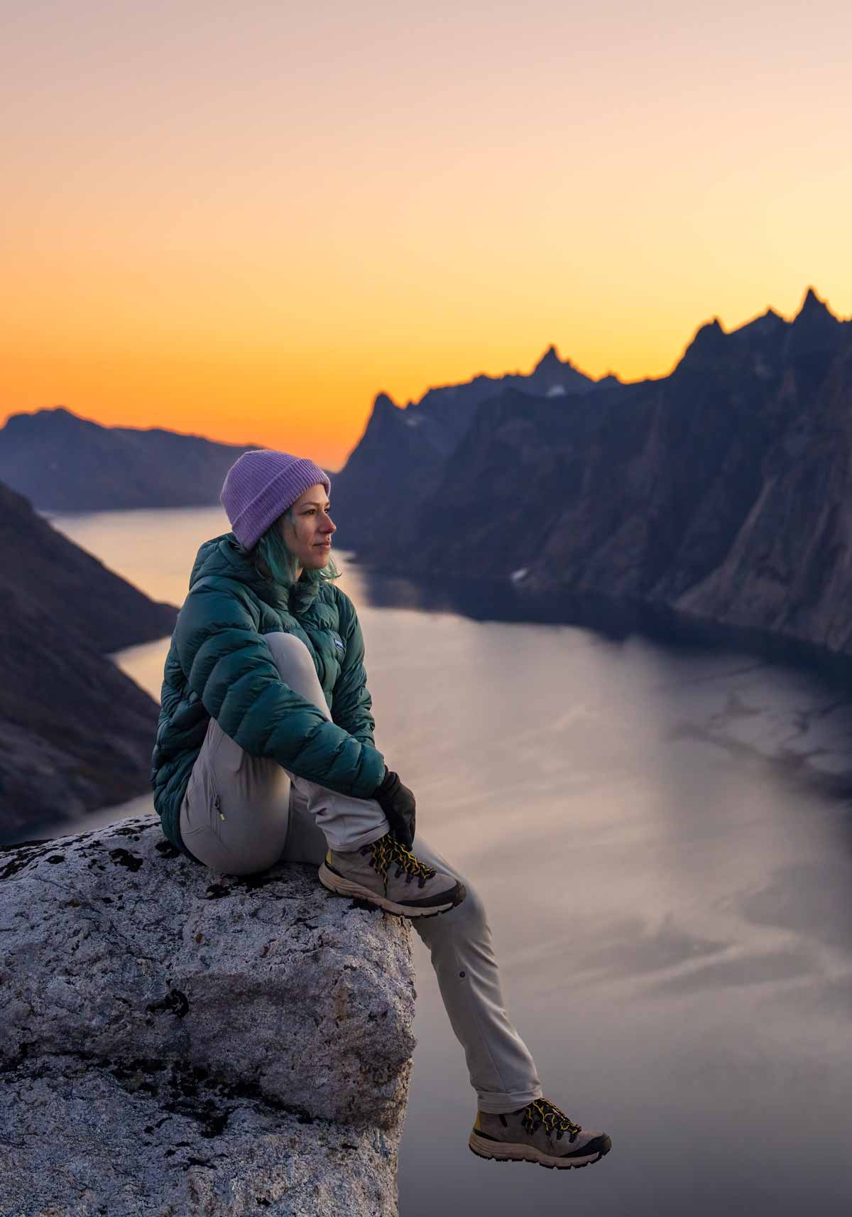 A woman wearing a Deep Teal Feathered Friends Eos Jacket sitting on a rock above a Greenland fjord at sunset