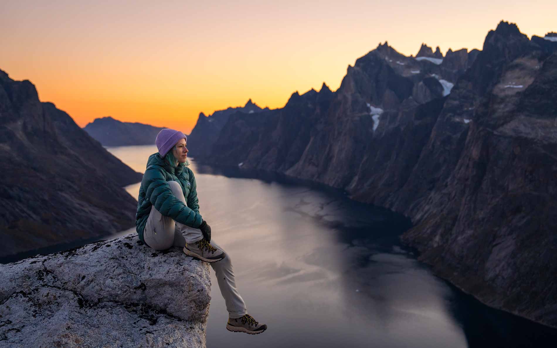 A woman wearing a Deep Teal Feathered Friends Eos Jacket sitting on a rock above a Greenland fjord at sunset landscape view