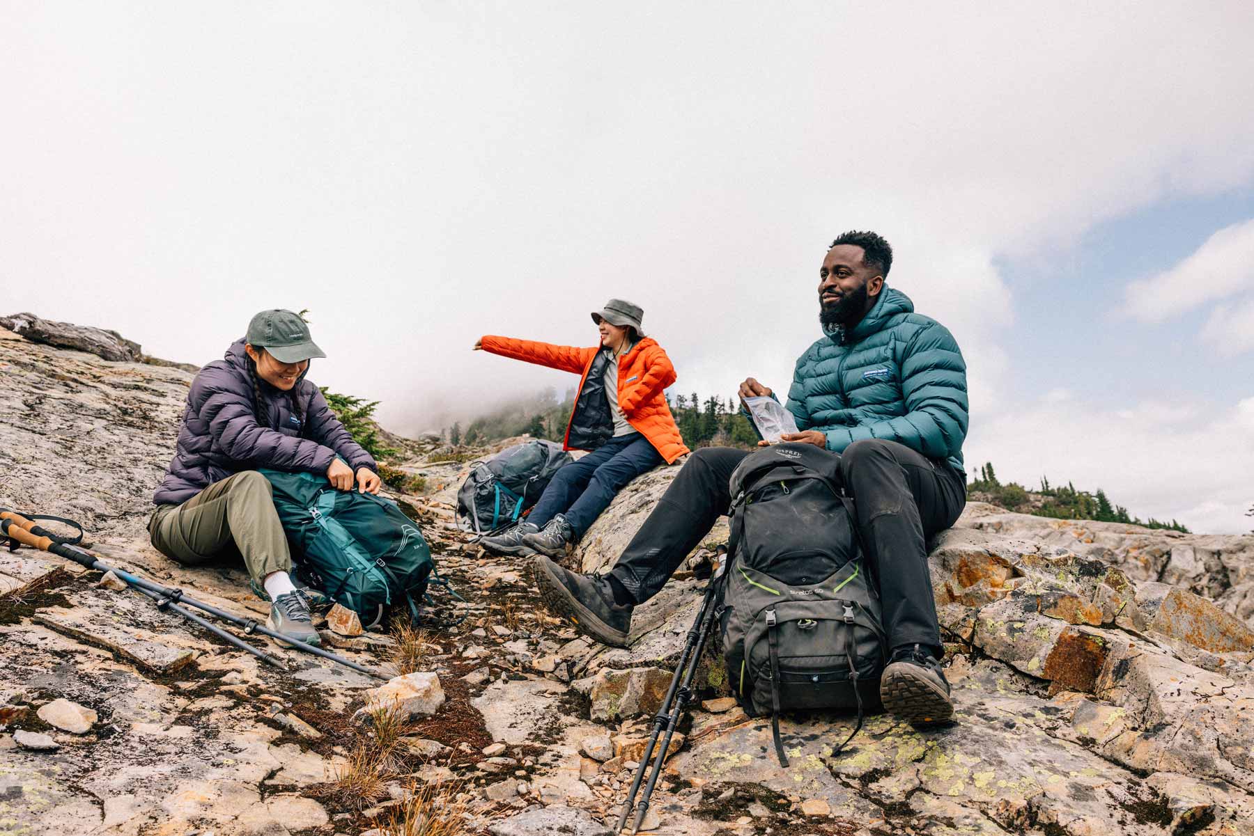 Two women and a man at a backpacking rest break wearing Feathered Friends Eos jackets