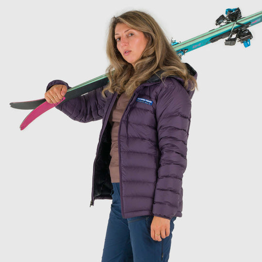 Studio front view of woman wearing a dusky purple Women's Feathered Friends Eos Down Jacket with skis on her shoulder