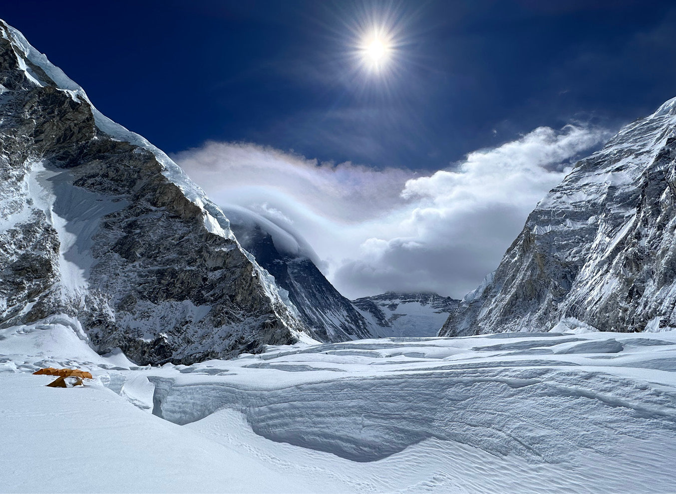 The sun sits high in a deep bluebird sky, framed by two cascading mountains cloaked in a veil of thick clouds. A little mustard-colored tent sits amongst a vast snowfield in the bottom left corner of a vast, white foreground.
