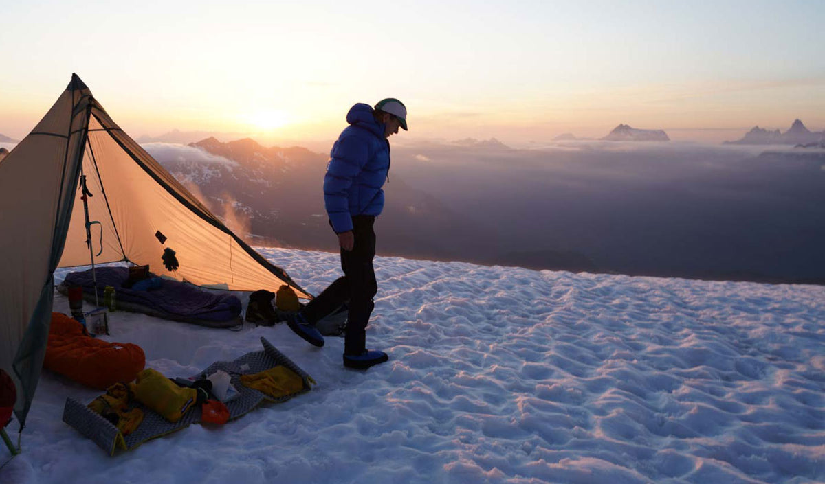 A camper wearing a blue down jacket beside a tent on a snowy ridge at sunset