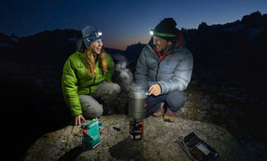 A man and woman wearing Feathered Friends Helios jackets cooking on a stove in the mountains after sunset