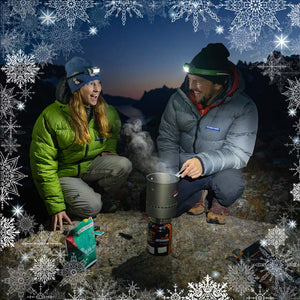 A woman wearing a Lichen Feathered Friends Helios jacket and a man wearing a gray Feathered Friends Helios down jacket cooking on a backpacking stove after sunset with a border of snowflakes