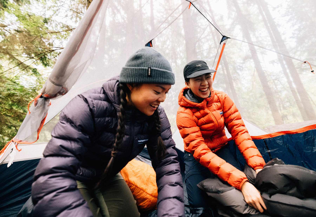 Two campers wearing purple and orange down jackets inside a tent in the forest