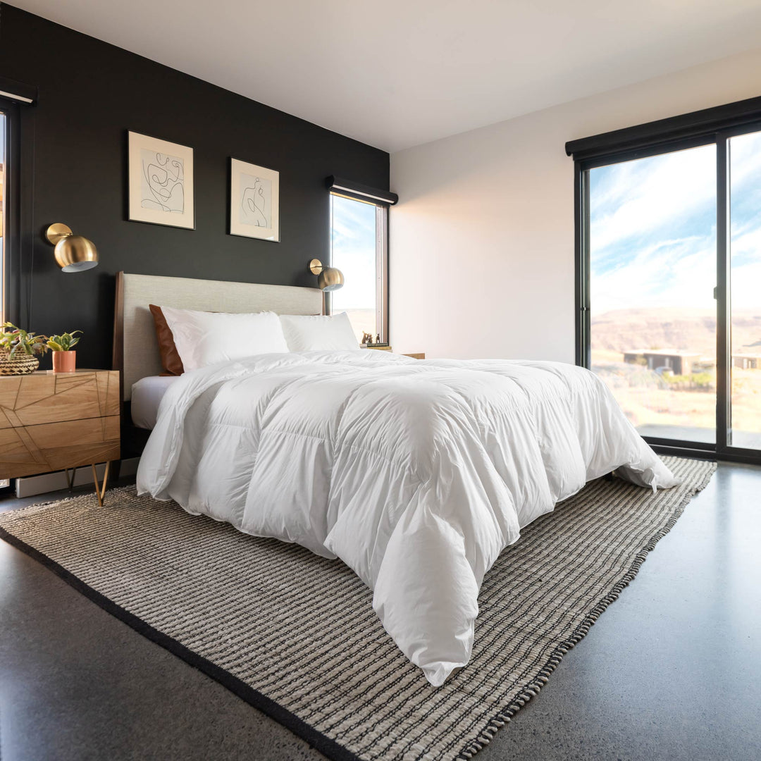 A modern bedroom featuring a white Feathered Friends Baffled Box 850 down comforter, set on a woven gray and black rug against a dark accent wall and desert landscape through the windows
