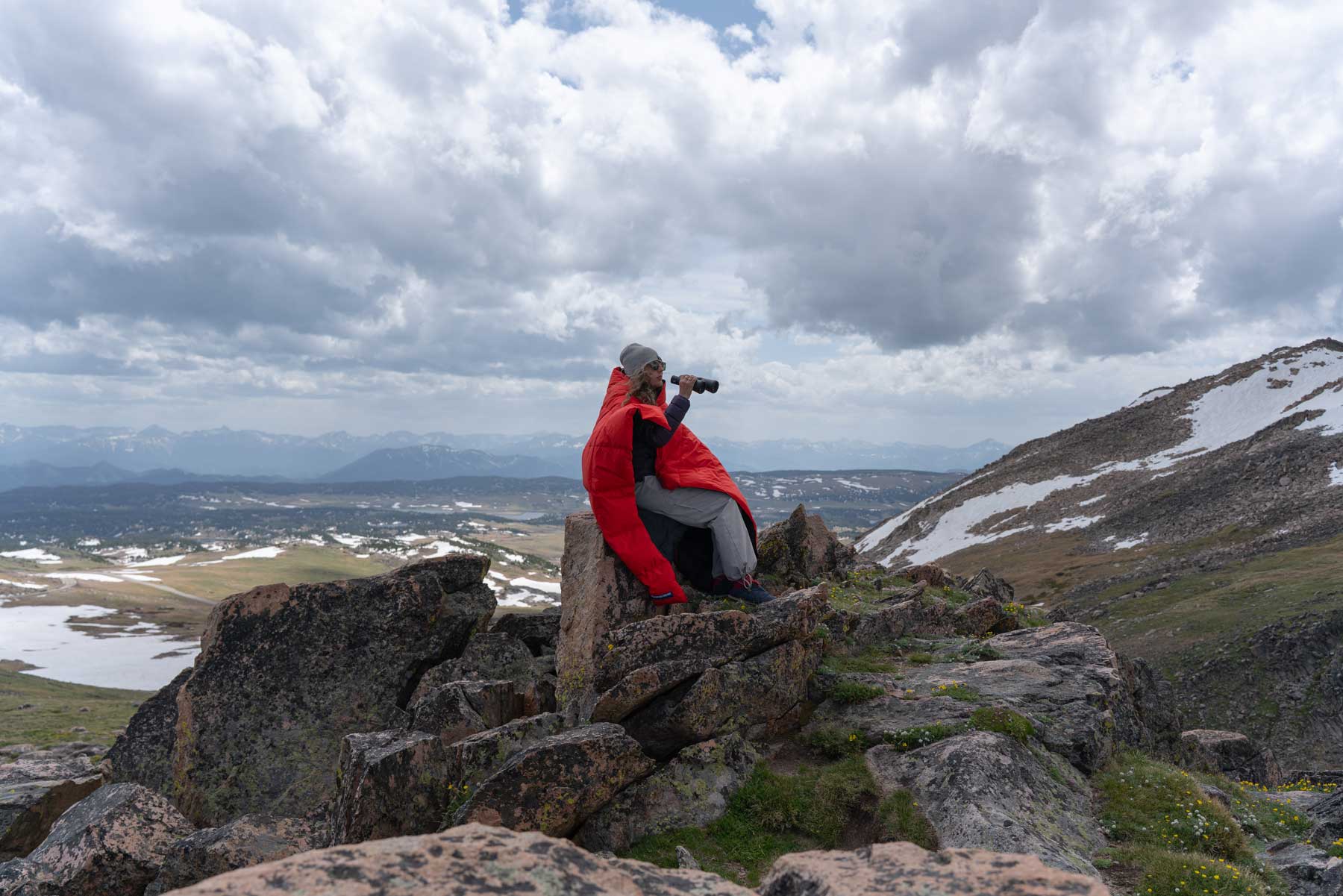 Wide open alpine landscape of a person sitting on a rock with binoculars draped in a red Feathered Friends down blanket