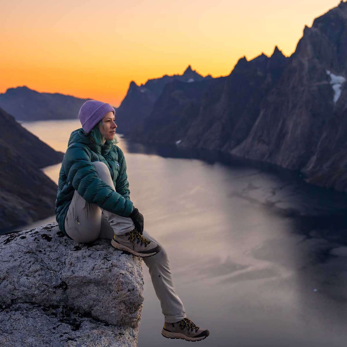 Woman sitting above a rocky fjord at sunset with a teal Eos Jacket