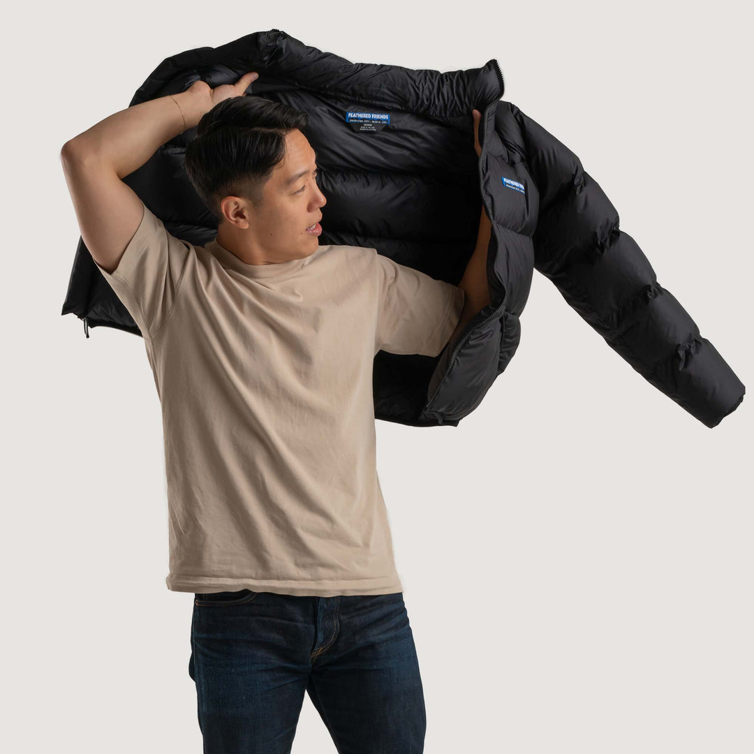 Studio side view of a man putting on a black Feathered Friends Helios Down Puffer jacket