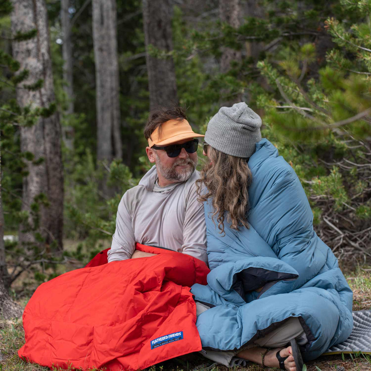 Two people sitting in a forest with a red and light blue Feathered Friends muscovy down blankets