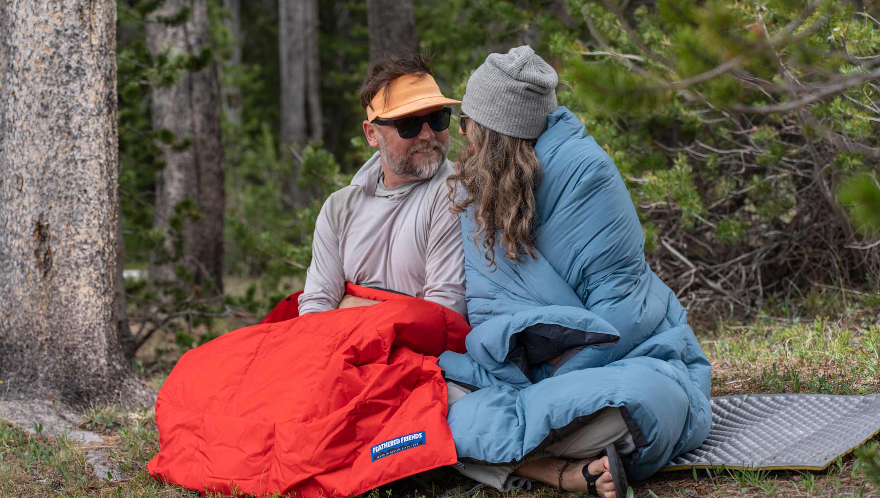 Wide view of two people sitting in a forest wrapped in red and light blue Feathered Friends down blankets