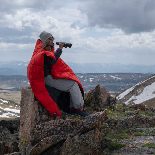 Person in a Feathered Friends water-resistant red down blanket sitting on a rocky outcrop with binoculars, looking over a mountainous landscape.