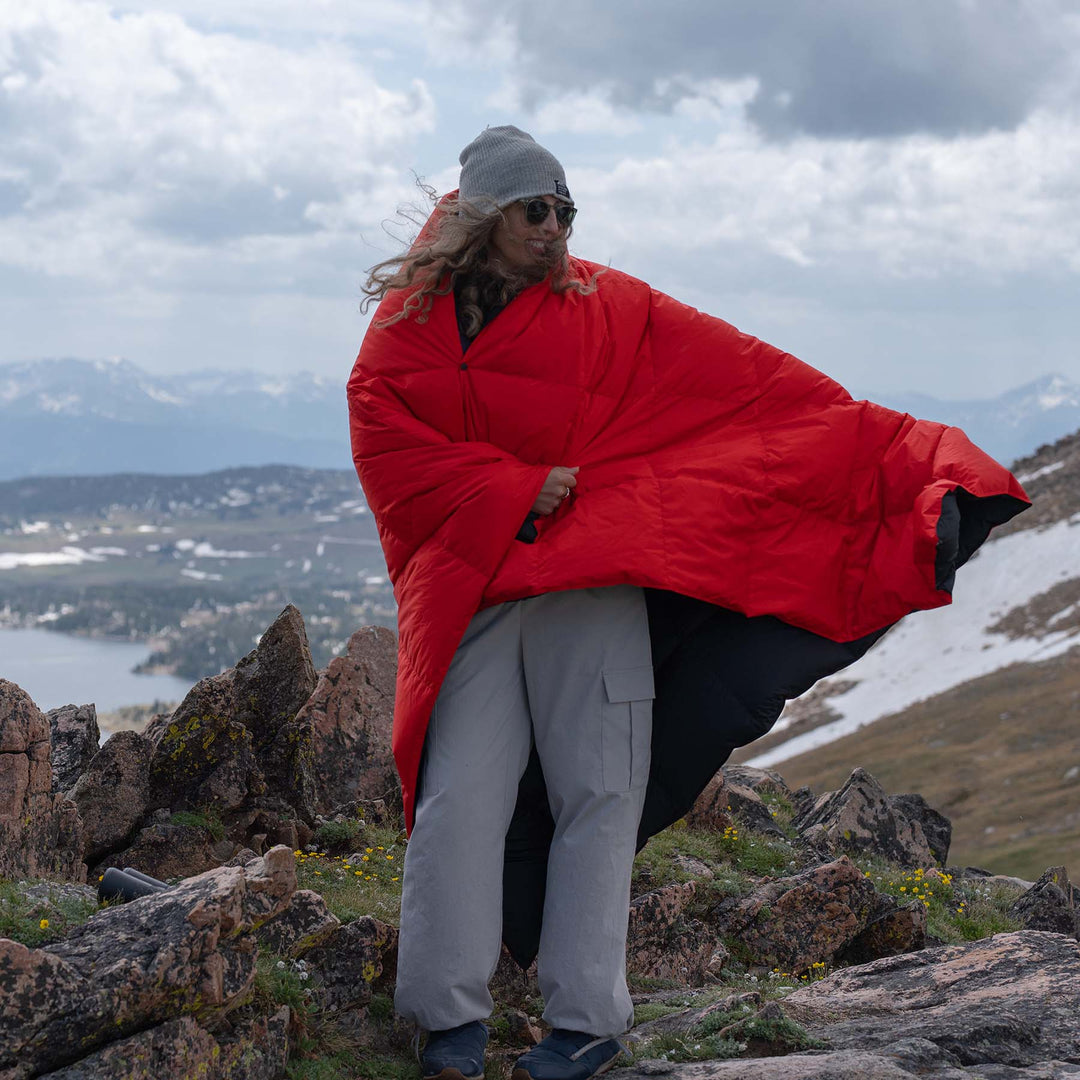 Person wrapped in a Feathered Friends red down blanket on a mountain with a scenic background