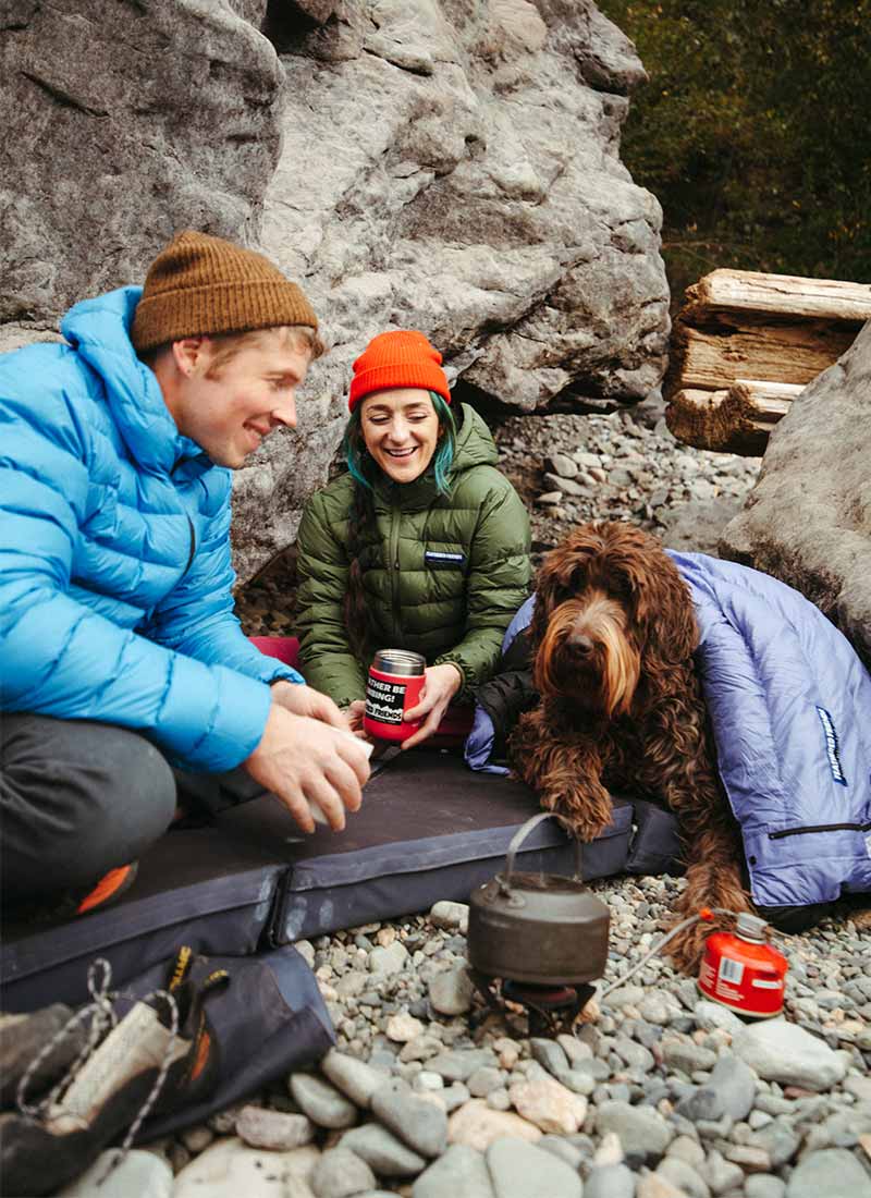 Man and woman sitting with Feathered Friends Eos Jackets and a sleeping bag over shaggy dog