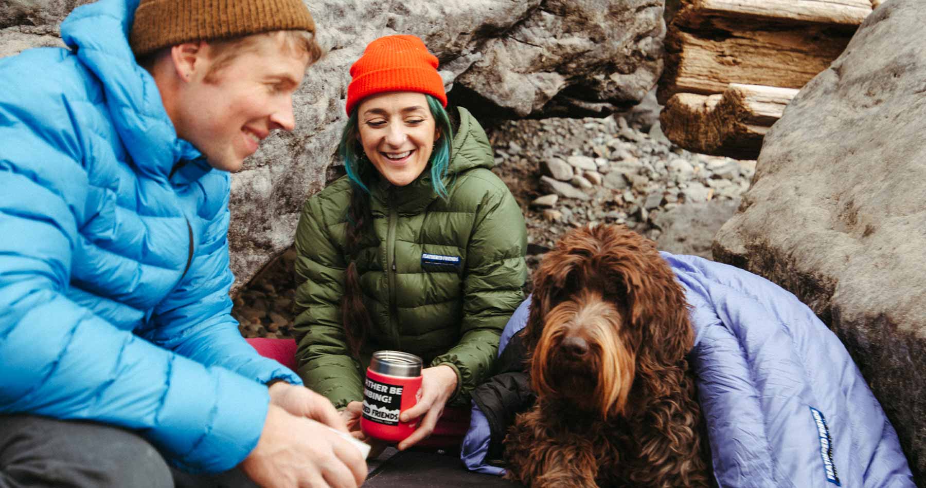 Man and woman sitting with Feathered Friends Eos Jackets and a sleeping bag over shaggy dog