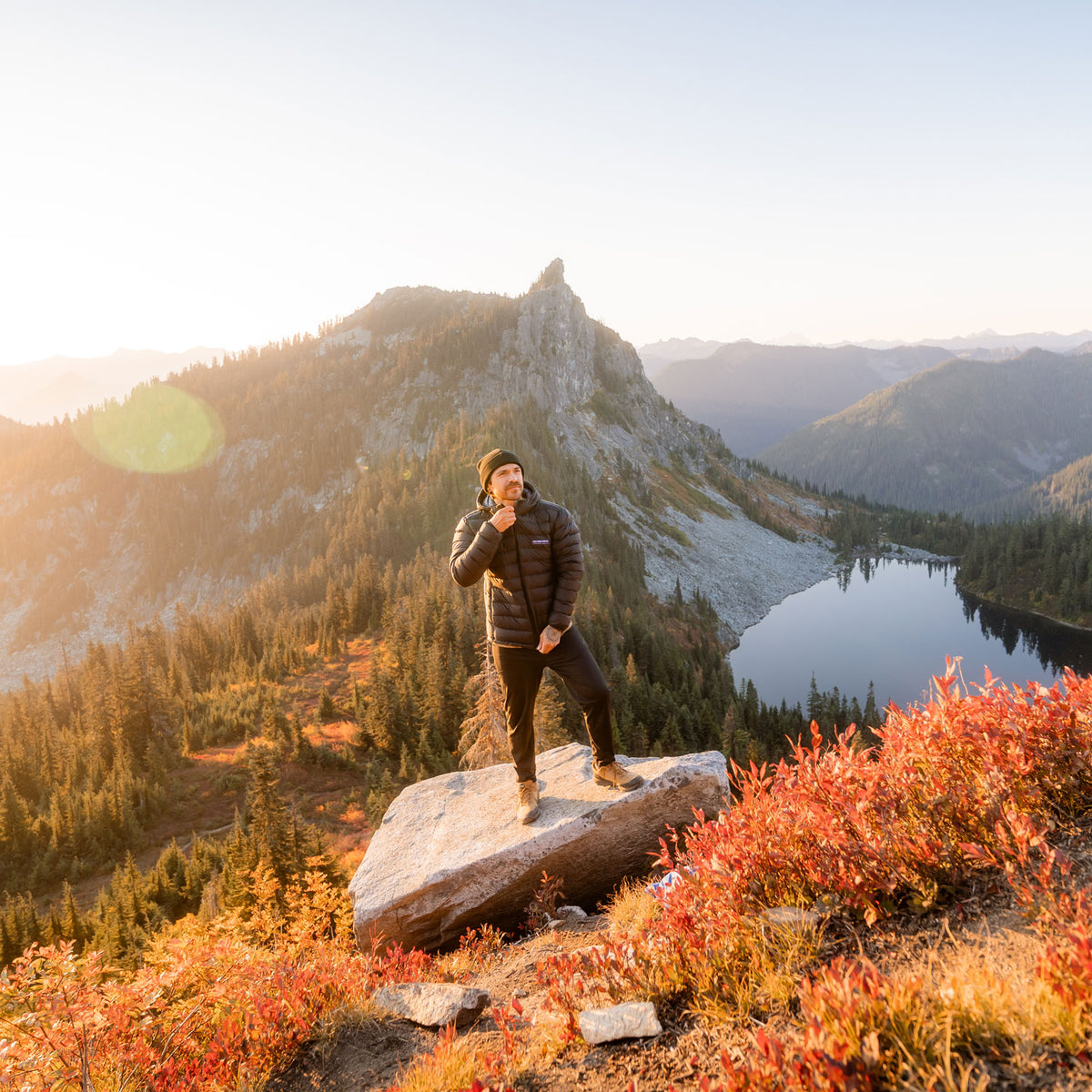 A man zipping up a black Feathered Friends Men's Eos Down Jacket on a granite boulder in the alpine during sunset with an alpine lake and peak in the background