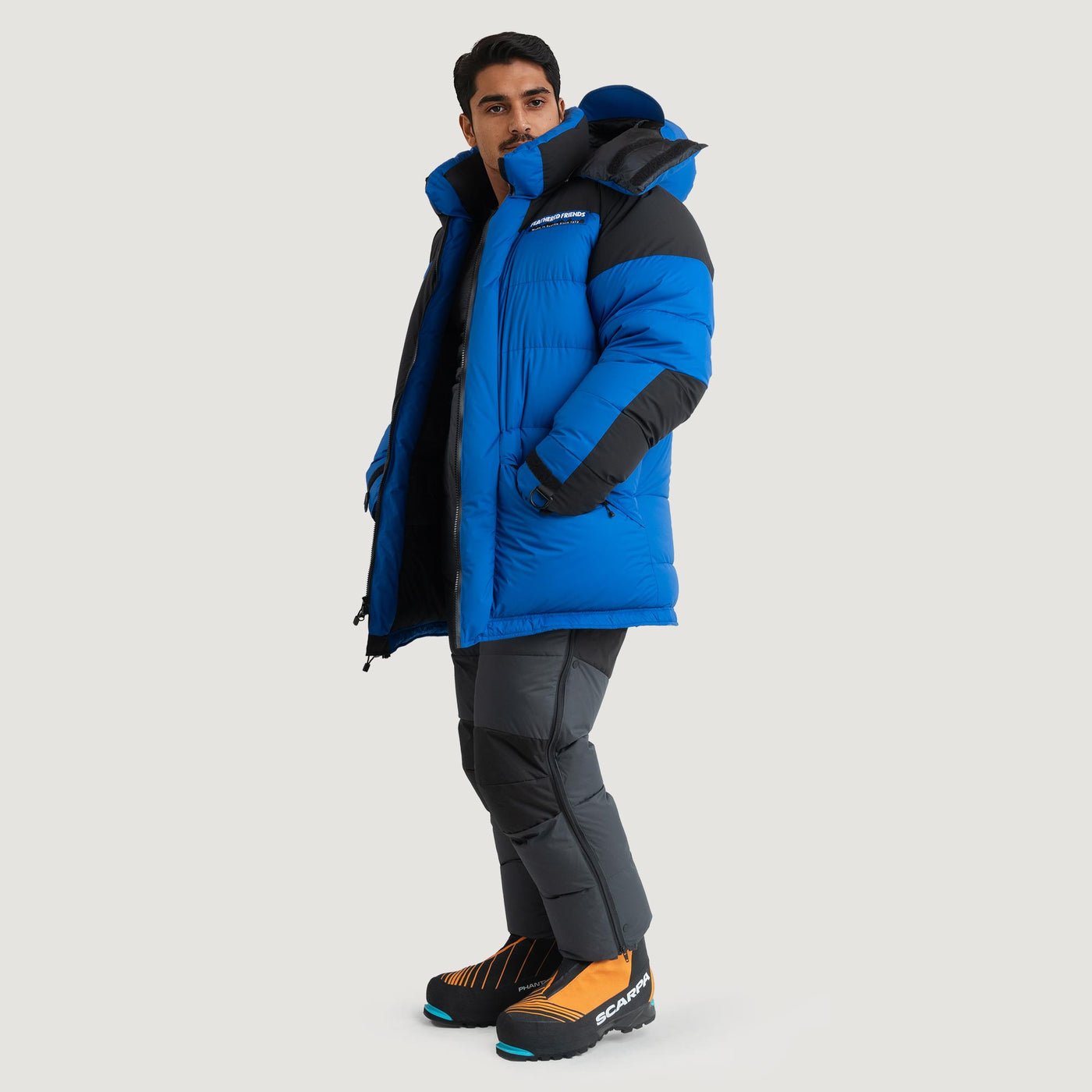 A model wearing the Feathered Friends Rock and Ice Expedition Parka in blue, wearing down pants and expedition climbing boots