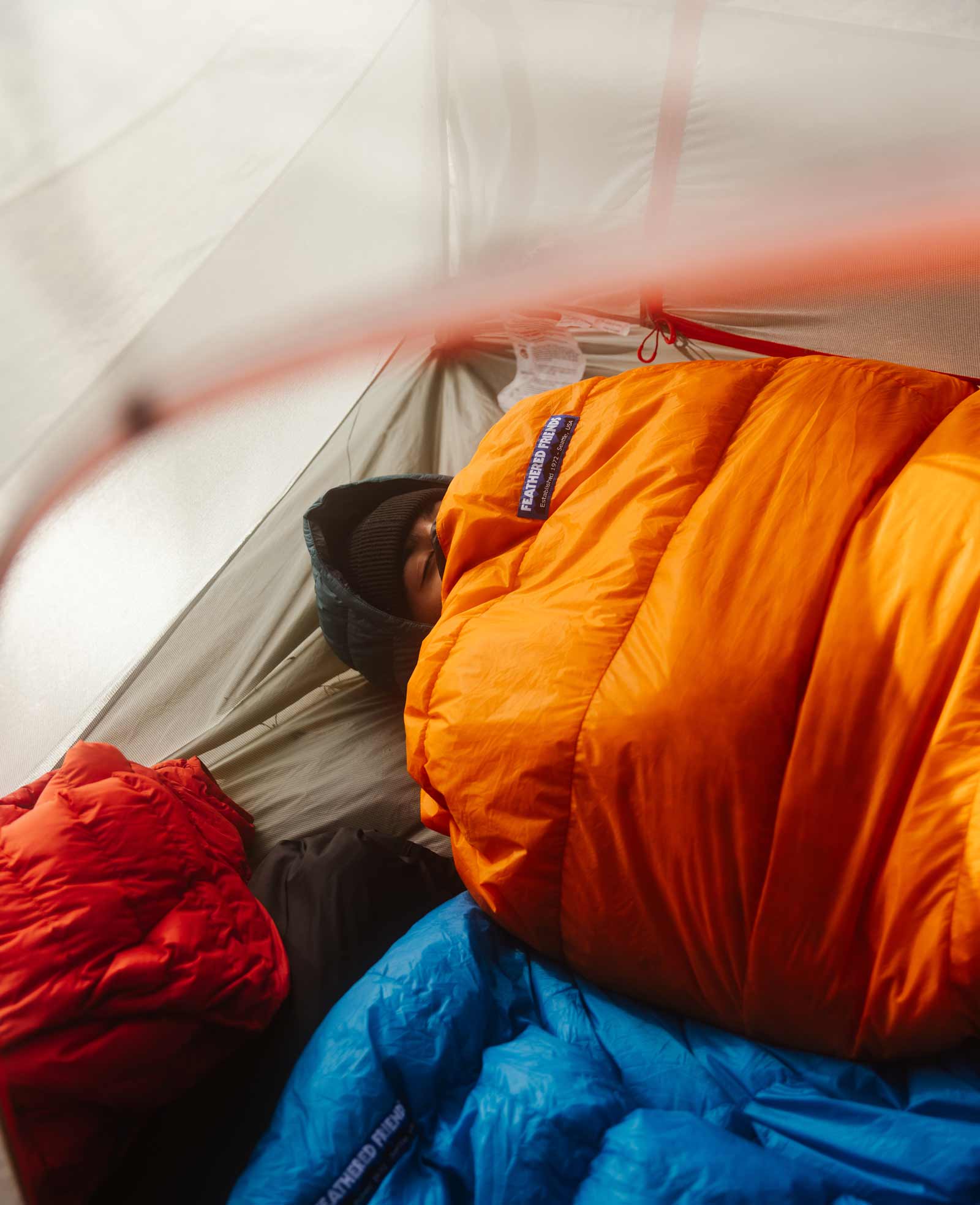 Person bundled in a flame orange Feathered Friends ultralight sleeping bag inside of a tent