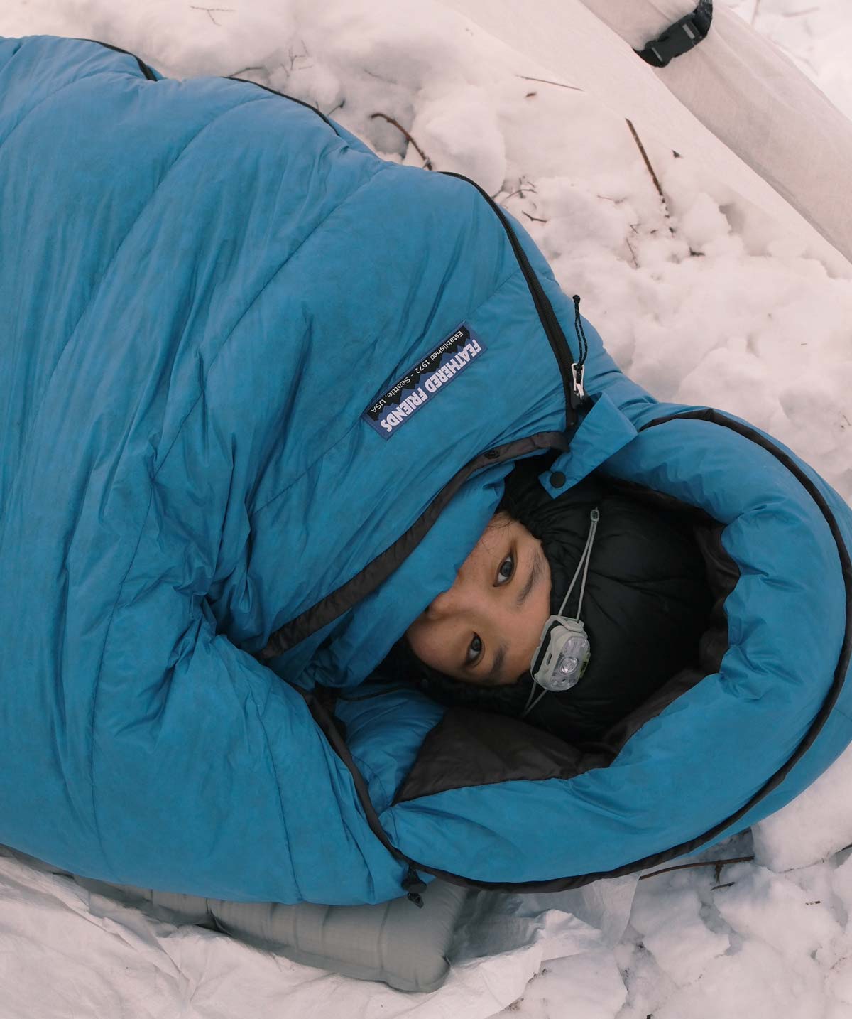 Person bundled in blue Winter Light Sleeping Bag on the snow with a headlamp on