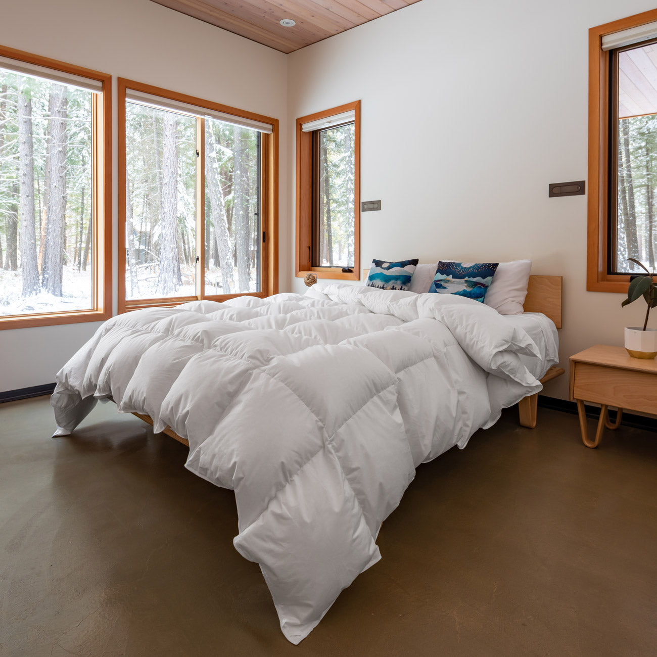 A white Feathered Friends Bavarian 850 down comforter on a bed with wooden frame, displayed in a bedroom with large windows showing a snowy landscape outside.
